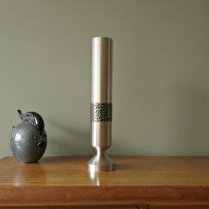 Könnte beinhalten: Eine hohe, silberfarbene Metallvase mit strukturiertem Band. Die Vase hat einen ausgestellten Sockel und einen zylindrischen Körper. Links von der Vase befindet sich eine dekorative Keramik-Schweinefigur. Auf einem Holzuntergrund.