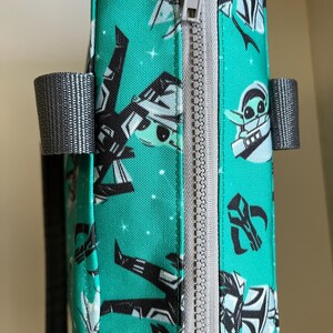 May include: A teal fabric bag featuring a repeating pattern of black and white cartoon characters and star motifs. The bag has a light gray zipper closure running vertically along the side and two gray fabric handles attached to the sides.