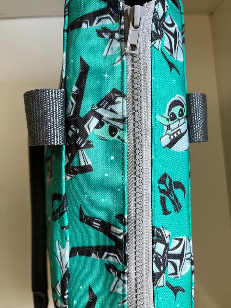 May include: A teal fabric bag featuring a repeating pattern of black and white cartoon characters and star motifs. The bag has a light gray zipper closure running vertically along the side and two gray fabric handles attached to the sides.