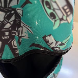 May include: A teal fabric bag featuring a repeating pattern of stylized characters in black, white, and light green. The bag has a black zipper closure at the top, revealing a black interior lining. The exterior fabric displays a space-themed design.