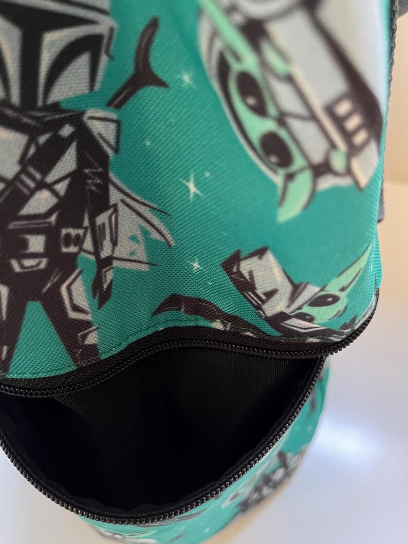 May include: A teal fabric bag featuring a repeating pattern of stylized characters in black, white, and light green. The bag has a black zipper closure at the top, revealing a black interior lining. The exterior fabric displays a space-themed design.