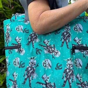 May include: A teal tote bag featuring a repeating pattern of cartoon characters in armor with small green creatures on their heads, accented by starburst shapes. The bag includes grey fabric straps and two front exterior pockets with black zippers.