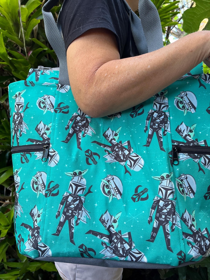 May include: A teal tote bag featuring a repeating pattern of cartoon characters in armor with small green creatures on their heads, accented by starburst shapes. The bag includes grey fabric straps and two front exterior pockets with black zippers.