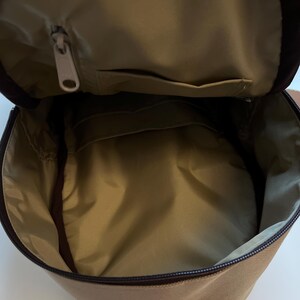 May include: A tan canvas backpack with a zippered top opening and a small pocket on the inside. The backpack has a brown zipper and brown straps.