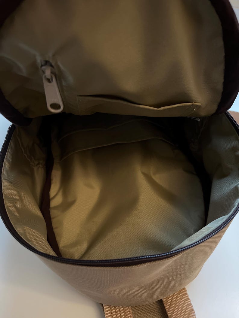 May include: A tan canvas backpack with a zippered top opening and a small pocket on the inside. The backpack has a brown zipper and brown straps.