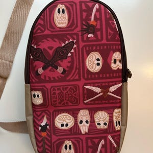 May include: A red and brown crossbody bag with a Polynesian pattern featuring tiki masks, spears, and geometric designs. The bag has an adjustable strap.