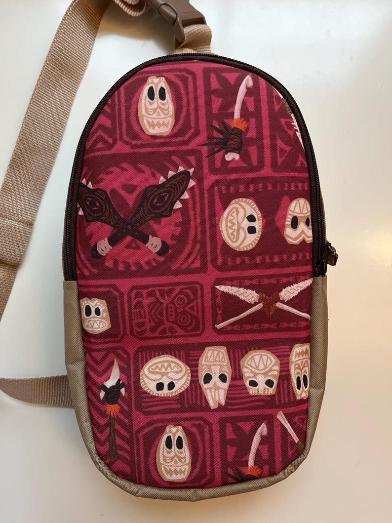 May include: A red and brown crossbody bag with a Polynesian pattern featuring tiki masks, spears, and geometric designs. The bag has an adjustable strap.