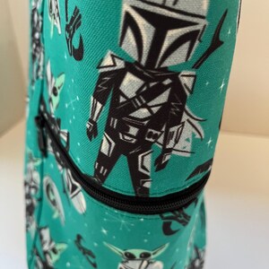 May include: A teal fabric bag featuring a repeating pattern of a cartoon armored character with a helmet and a small green creature. The bag includes a black zipper closure that runs horizontally across the middle of the cylindrical structure.