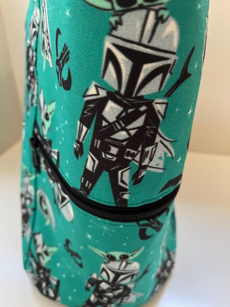 May include: A teal fabric bag featuring a repeating pattern of a cartoon armored character with a helmet and a small green creature. The bag includes a black zipper closure that runs horizontally across the middle of the cylindrical structure.