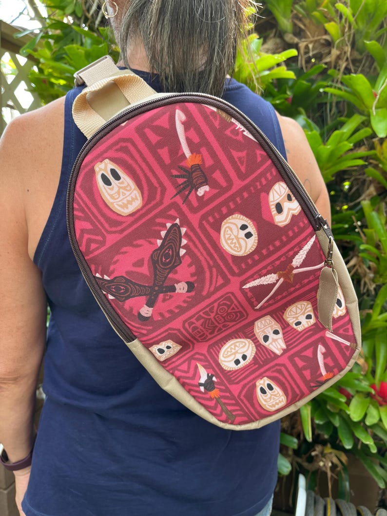 May include: A red and tan crossbody bag with a Polynesian-inspired print featuring tiki masks, spears, and other tribal designs. The bag has a zipper closure and a long adjustable strap.