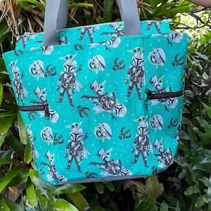 May include: A teal tote bag featuring a repeating pattern of cartoon characters in armor and small green creatures, accented with gray fabric handles and black zippered side pockets. The design includes small white star-like shapes across the fabric.