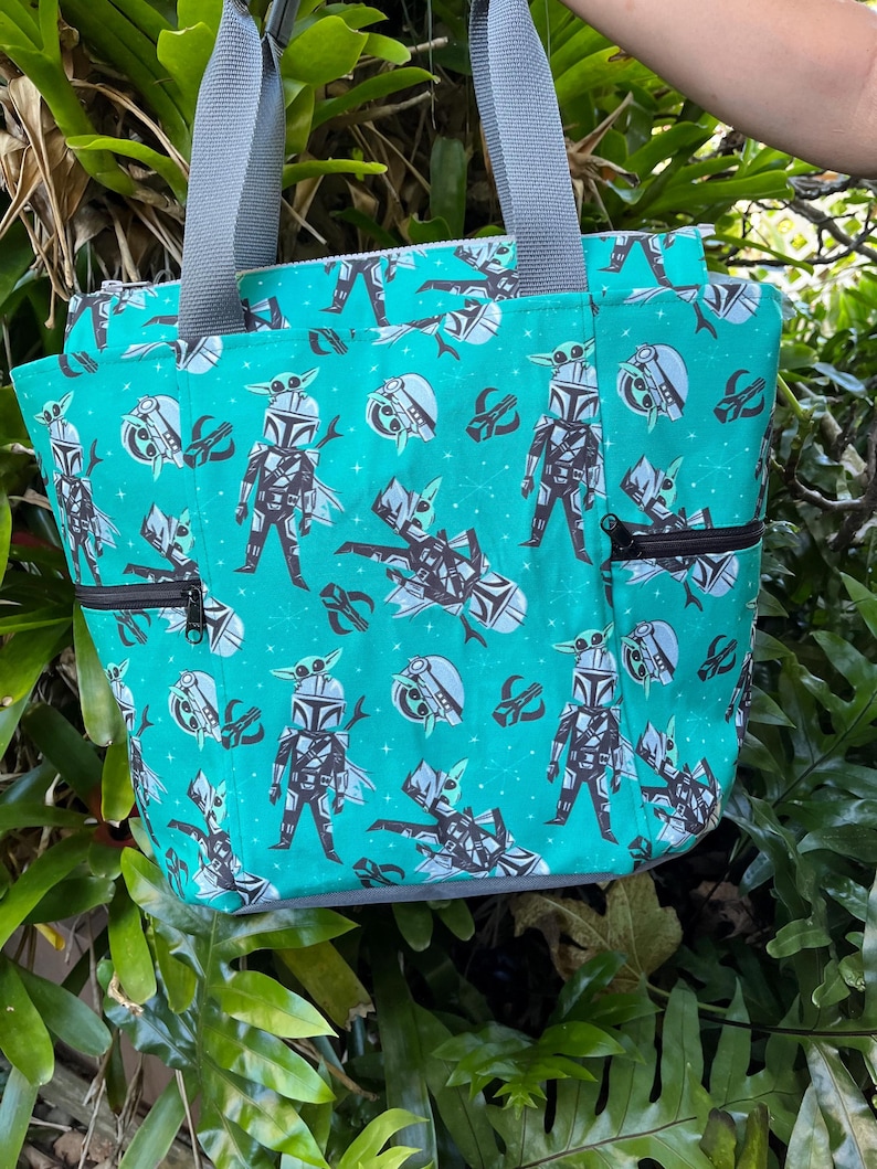 May include: A teal tote bag featuring a repeating pattern of cartoon characters in armor and small green creatures, accented with gray fabric handles and black zippered side pockets. The design includes small white star-like shapes across the fabric.
