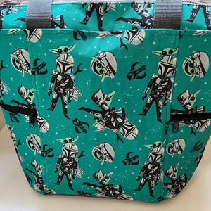 May include: A teal tote bag featuring a repeating pattern of a cartoon armored character with a small green creature on their head. The bag includes grey fabric handles, a top zipper closure, and two front pockets with black zippers.