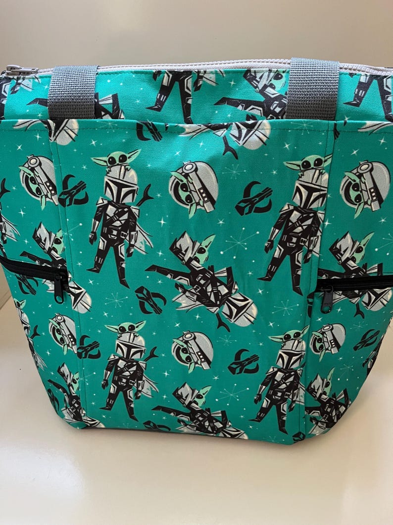 May include: A teal tote bag featuring a repeating pattern of a cartoon armored character with a small green creature on their head. The bag includes grey fabric handles, a top zipper closure, and two front pockets with black zippers.