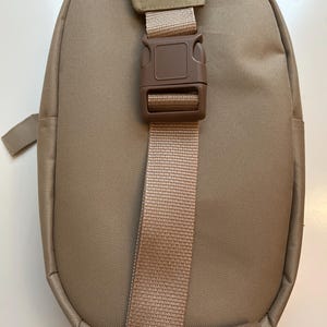 May include: A tan-colored backpack with a single adjustable strap and a brown plastic buckle. The backpack has a rounded shape and a smooth fabric texture.