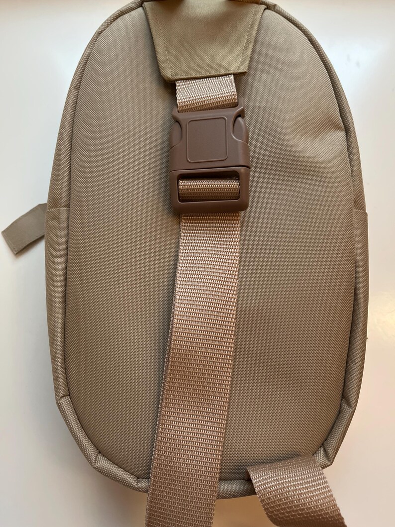 May include: A tan-colored backpack with a single adjustable strap and a brown plastic buckle. The backpack has a rounded shape and a smooth fabric texture.