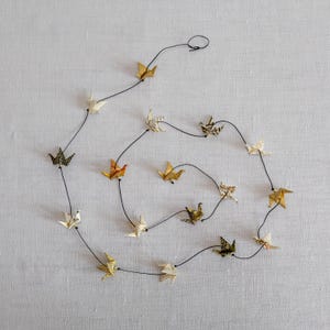 Pode incluir: Uma guirlanda de origamis de papel em várias cores, incluindo dourado, branco e estampas, enfiados em fio preto. Os origamis estão dispostos em um padrão circular e fluido sobre um fundo cinza claro.