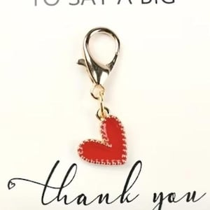May include: A gold-colored lobster clasp charm with a red heart pendant. The heart has a stitched border design. The image includes the text "A LITTLE HUG TO SAY A BIG thank you" in black script.