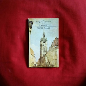 Puede incluir: Un libro de bolsillo de Penguin Classics titulado "Flaubert Three Tales". La portada presenta una ilustración detallada de una ciudad europea con una torre del reloj. El libro está sobre un fondo rojo.