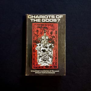 May include: A book titled "Chariots of the Gods?" with a black and red cover. The cover displays a detailed illustration of a figure with intricate designs. The book's subtitle reads "Unsolved mysteries of the past" by Erich Von Daniken.