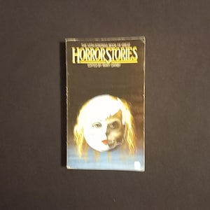 以下が含まれることがあります： メアリー・ダンビー編集の「Horror Stories」というタイトルのヴィンテージペーパーバック。表紙には、半分が白く、もう半分が朽ち果てた骸骨のような外観の顔が描かれています。タイトルは太字の黄色文字です。