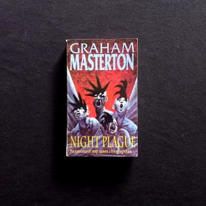 Op de afbeelding: Een paperback boekomslag met een rode, zwarte en witte illustratie van drie figuren met stekelig haar. De titel van het boek is "Night Plague" en de auteur is "Graham Masterton". De tekst onder de titel luidt "The innocence of sleep spawns a living nightmare..."