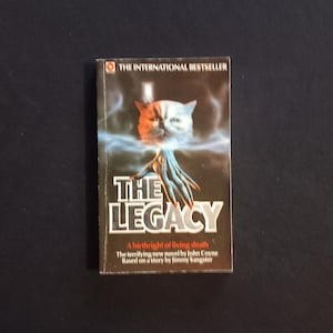 Może przedstawiać: Książka w miękkiej oprawie zatytułowana "The Legacy" z surrealistyczną okładką przedstawiającą stworzenie przypominające kota z niebieskimi rękami i ognistą pomarańczowo-białą głową. Książka jest oznaczona jako "The International Bestseller".