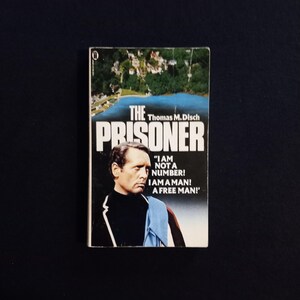 May include: A vintage paperback book titled "The Prisoner" by Thomas M. Disch. The cover features a man's portrait and the text "I AM NOT A NUMBER! I AM A MAN! A FREE MAN!". The background shows a coastal landscape.