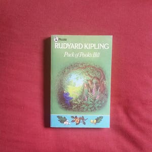 Puede incluir: Un libro de bolsillo verde con el título "Puck of Pook's Hill" de Rudyard Kipling. La portada presenta una ilustración de una escena de bosque con dos niños y una figura azul.