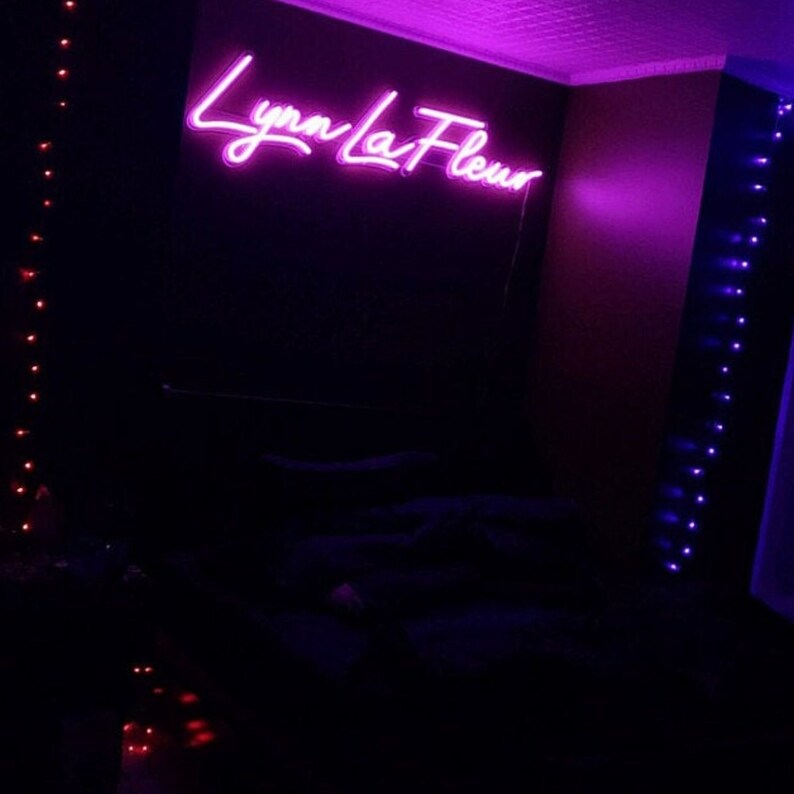 Puede incluir: Un dormitorio con un letrero de ne&oacute;n que dice "Lynn La Fleur" en color morado oscuro. La habitaci&oacute;n est&aacute; iluminada con luces de cadena moradas y rojas.