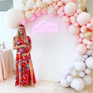 Puede incluir: Una mujer embarazada con un vestido maxi floral se encuentra frente a una pared blanca con un letrero de ne&oacute;n rosa que dice "Aria" y un arco de globos hecho de globos rosas, blancos y dorados.