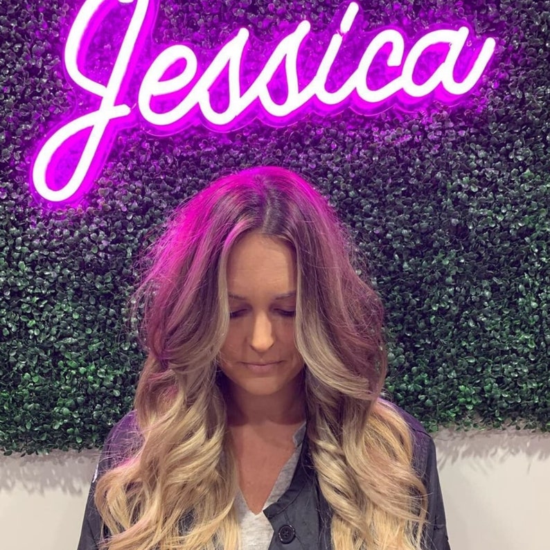Puede incluir: Una mujer con el pelo largo y rubio, peinado con rizos sueltos, se encuentra frente a un letrero de ne&oacute;n morado que dice "Jessica". El letrero est&aacute; en una pared cubierta de c&eacute;sped artificial verde.