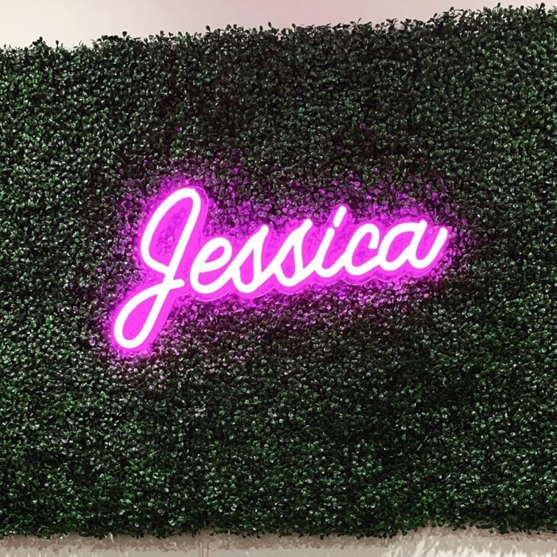 Puede incluir: Un letrero de ne&oacute;n rosa con el nombre "Jessica" en una pared verde de follaje artificial.