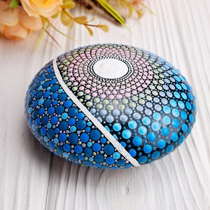 May include: A hand-painted rock with a blue, green, and pink mandala design. The rock is painted with small dots and has a white line dividing the design in half.