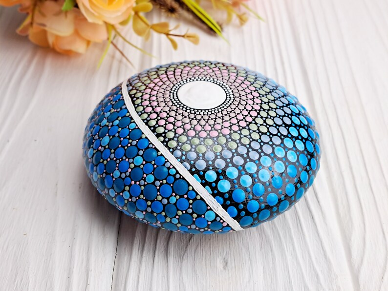 May include: A hand-painted rock with a blue, green, and pink mandala design. The rock is painted with small dots and has a white line dividing the design in half.