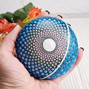 May include: A hand holds a round, painted rock with a mandala design. The mandala features a white center surrounded by concentric circles of pink, green, and blue dots. The rock is painted blue on the other side.