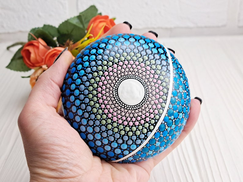 May include: A hand holds a round, painted rock with a mandala design. The mandala features a white center surrounded by concentric circles of pink, green, and blue dots. The rock is painted blue on the other side.
