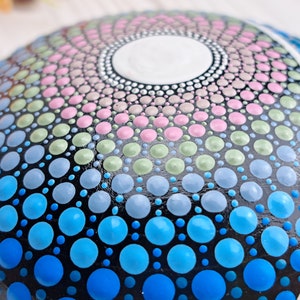 May include: Close-up of a painted rock with a mandala design. The design features a circular pattern of dots in shades of blue, green, pink, and white on a black background.