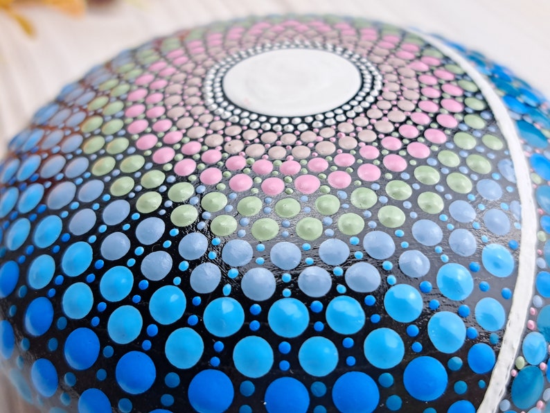 May include: Close-up of a painted rock with a mandala design. The design features a circular pattern of dots in shades of blue, green, pink, and white on a black background.