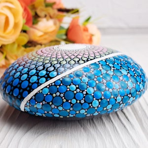 May include: A round, painted rock with a mandala design in shades of blue, pink, and green. The rock is painted with small dots and has a white line dividing the design in half.