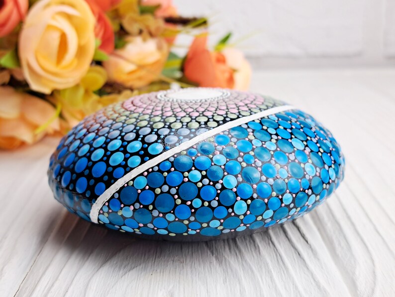 May include: A round, painted rock with a mandala design in shades of blue, pink, and green. The rock is painted with small dots and has a white line dividing the design in half.