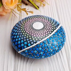 May include: A round, painted rock with a mandala design in shades of blue, green, pink, and white. The rock is painted with dots of various sizes.
