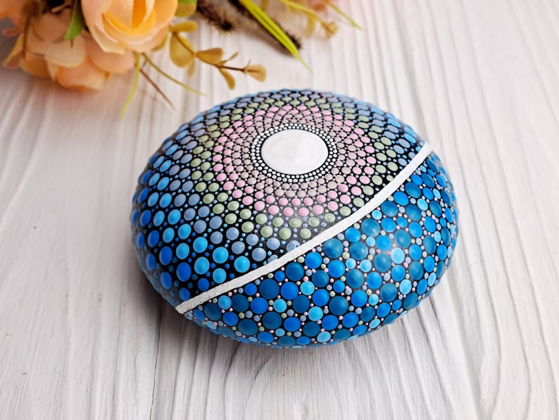 May include: A round, painted rock with a mandala design in shades of blue, green, pink, and white. The rock is painted with dots of various sizes.