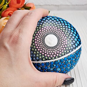 May include: A hand holds a round, painted rock with a mandala design. The mandala features a white center circle surrounded by concentric circles of pink, green, and blue dots. The bottom half of the rock is painted in shades of blue.