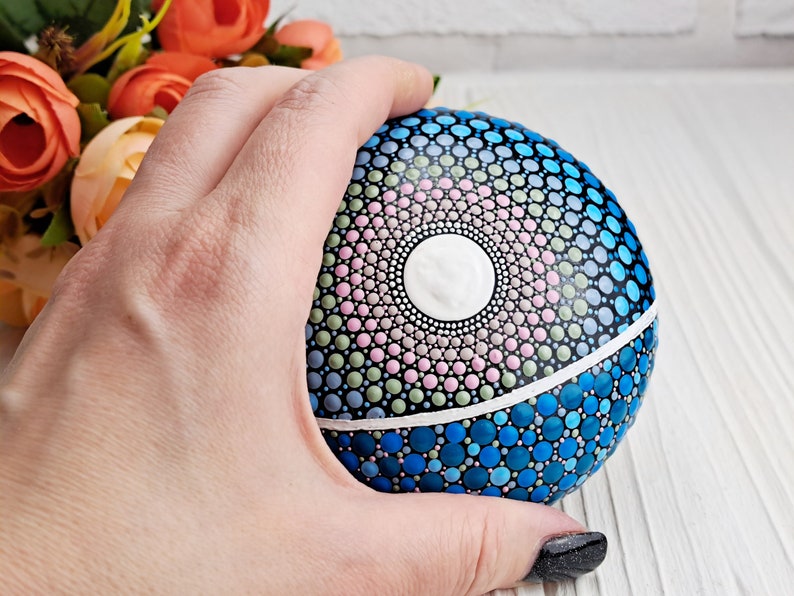May include: A hand holds a round, painted rock with a mandala design. The mandala features a white center circle surrounded by concentric circles of pink, green, and blue dots. The bottom half of the rock is painted in shades of blue.