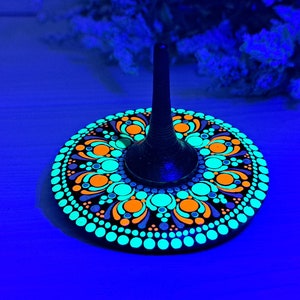 May include: A black spinning top with a colorful mandala design painted on the base. The design features orange, green, and blue dots and swirls on a black background. The top is illuminated with a blacklight, making the colors glow brightly.