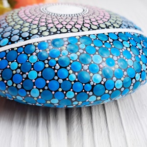 May include: A round, smooth stone painted with a mandala design in shades of blue, pink, and green. The stone is painted with small dots and circles, creating a detailed and intricate pattern.