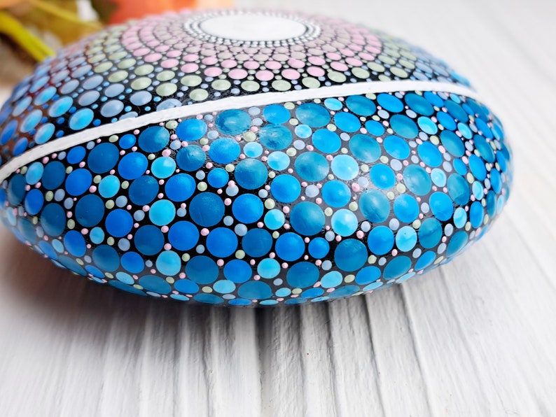 May include: A round, smooth stone painted with a mandala design in shades of blue, pink, and green. The stone is painted with small dots and circles, creating a detailed and intricate pattern.