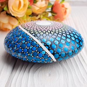 May include: A hand-painted rock with a mandala design in shades of blue, pink, and green. The rock is painted with small dots and has a white line dividing the design.