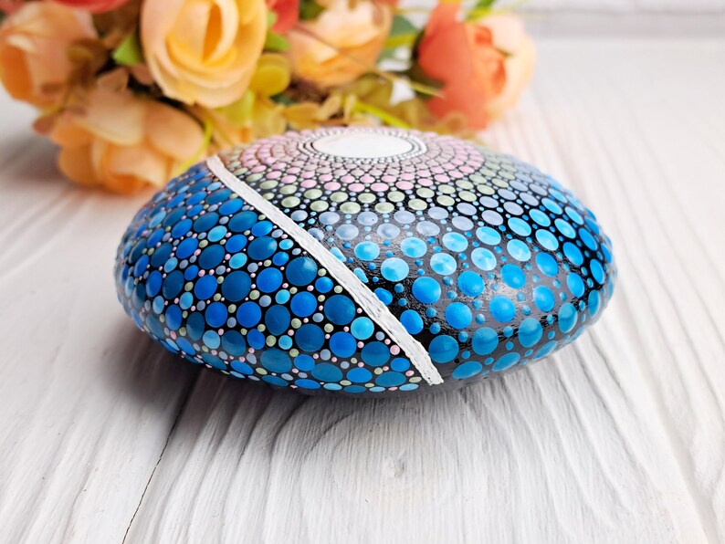 May include: A hand-painted rock with a mandala design in shades of blue, pink, and green. The rock is painted with small dots and has a white line dividing the design.